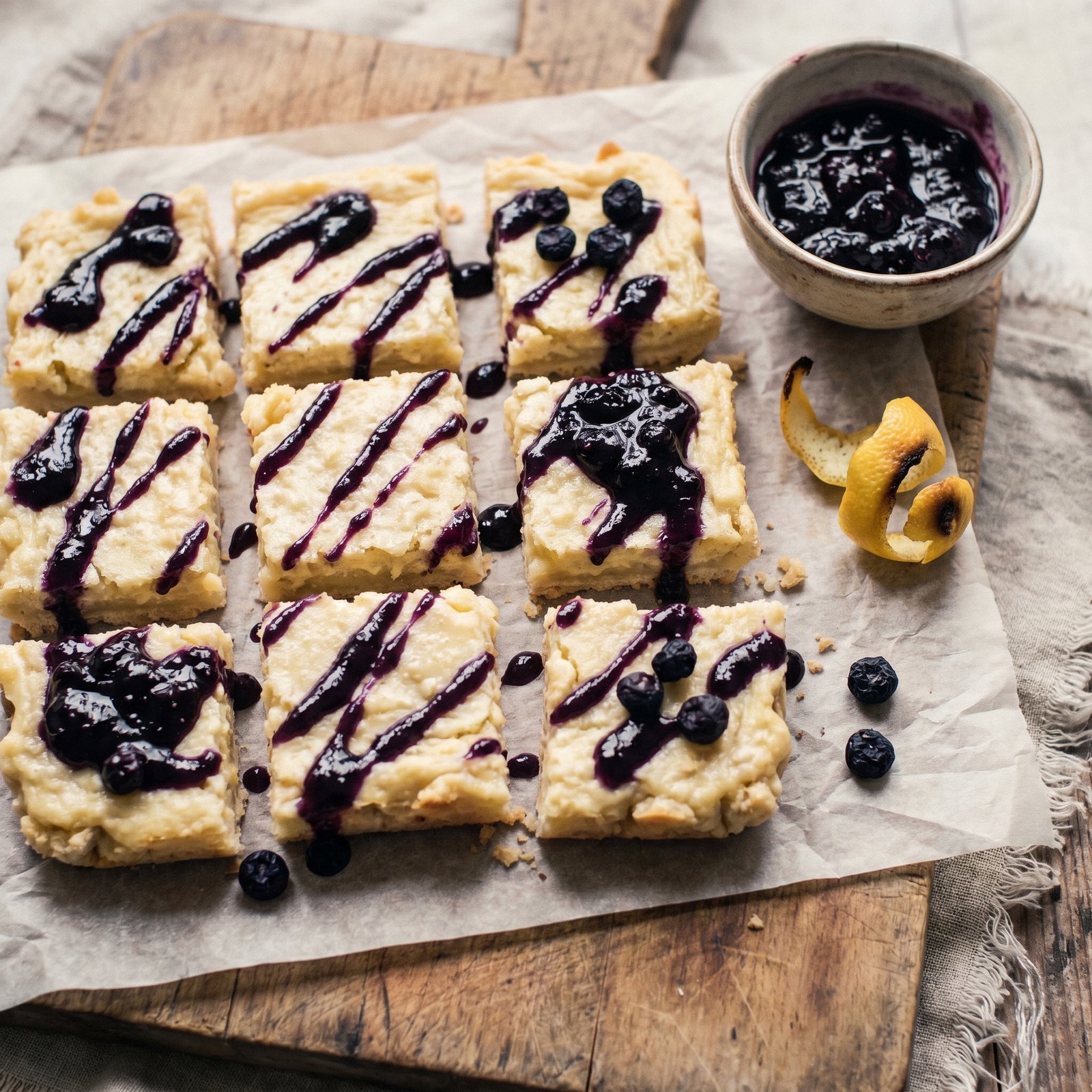 Nine pale-yellow lemon icebox bars in a grid on parchment paper, each drizzled with a glossy indigo blueberry preserve ribbon, on pale marble.
